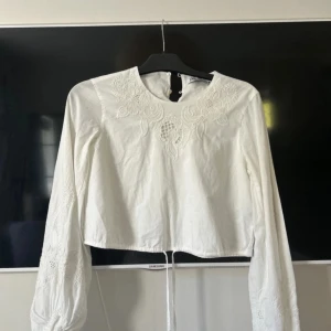 Vit broderad blus från Zara - Superfin vit blus från Zara med broderade blommor på bröstet och ärmarna. Långärmad modell med knäppning i nacken och croppad passform. Perfekt för dig som gillar romantiska detaljer och stilren look. Endast testad
