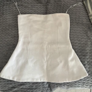Vit korsett-topp i satin - Säljer en snygg vit korsett-topp med utsvängd nederdel. Toppen har en stilren och minimalistisk look med släta sömmar framtill. Materialet är glansigt satin som ger en lyxig känsla och passar perfekt till trendiga outfits.