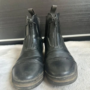 Svarta ridskor med dragkedja - Säljer ett par svarta ridskor från CRW i skinn med dragkedja framtill och elastiska paneler på sidorna. De har rund tå, platt sula och praktiska flikar bak för enkel påtagning. Perfekta för dig som gillar stilren och funktionell look. 