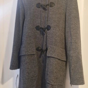 Grå duffelcoat med huva från Classic - Säljer en grå duffelcoat med huva från Classic. Jackan har stora fickor framtill och snygga svarta plastknappar med öglor. Tillverkad i ullblandning med mjukt foder, perfekt för kyliga dagar. Klassisk och tidlös modell som håller dig varm hela vintern.