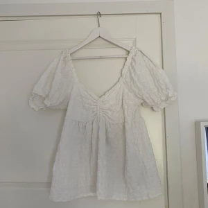 Vit blus med puffärmar och v-ringning - Supersöt vit blus med puffiga korta ärmar och v-ringning framtill. Toppen har rynkade detaljer vid bysten och är tillverkad i ett lätt, strukturerat tyg som ger en romantisk känsla. Perfekt för dig som gillar en luftig och feminin stil.