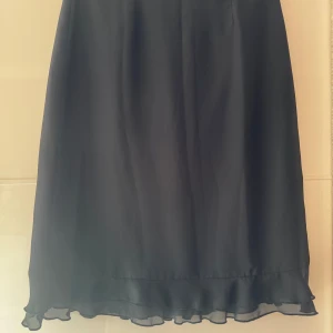 Svart kjol med volang från Lindex - Svart kjol från Lindex i 100% polyester. Kjolen har en rak passform och är dekorerad med en snygg volang längst ner som ger en extra touch. Perfekt för dig som gillar stilrena plagg med en liten twist. Storleklapp avklippt, men den är L, midjan 84cm.