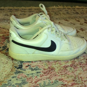 Nike vita sneakers med svart swoosh - Fräscha vita sneakers från Nike med klassisk svart swoosh på sidan. Modellen har snörning, perforerad tå och platt sula. Skorna är låga och tillverkade i skinn med en mjuk insida för extra komfort. Perfekta för dig som gillar stilrena och sportiga sneakers.