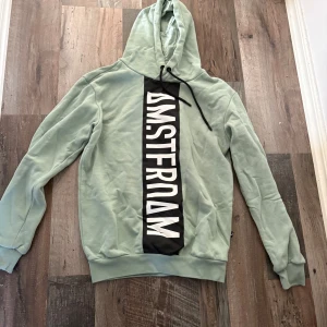 Mintgrön hoodie med tryck - Snygg mintgrön hoodie med svart och vitt tryck framtill där det står 'AMSTERDAM' i stora bokstäver. Tröjan har huva med snörning och långa ärmar. Perfekt för dig som gillar streetstyle och vill sticka ut lite extra.