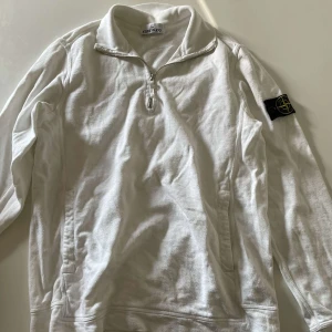 Vit half zip tröja från Stone Island - Vit långärmad tröja från Stone Island med klassisk patch på ärmen. Tröjan har en snygg half zip-dragkedja vid kragen och är tillverkad i mjuk bomull. Perfekt för dig som gillar stilrena och sportiga plagg med premiumkänsla.