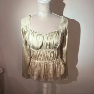 Champagnefärgad blus i satinlook - Supersöt blus i champagnefärgad satinlook med långa ärmar och markerad midja. Toppen har en lätt volang nertill och smockade detaljer som ger en romantisk vibe. Perfekt för dig som gillar en feminin och trendig stil.