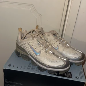 Nike Air Vapormax beige sneakers - Snygga Nike Air Vapormax sneakers i beige med silvriga detaljer och reflekterande Swoosh-logga. Skorna har en futuristisk design med genomskinlig bubblig sula och snörning. Perfekta för dig som vill sticka ut med en modern och sportig look.