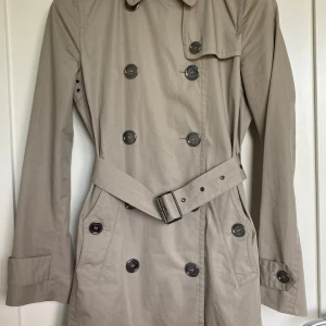 Burberry Brit trenchcoat  - Klassikern Burberry Brit - stilren trenchcoat med dubbelknäppning och bälte i midjan. Klassisk krage med stora knappar framtill. Som ny. Perfekt för dig som vill ha en tidlös och snygg kappa. Kan även mötas i centrala- eller norra Stockholm för inbetalning och överlämning. 