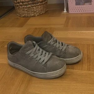 Grå mockasneakers från Selected Homme - Säljer ett par stilrena grå sneakers från Selected Homme i mocka. Skorna har rund tå, platt sula och klassisk snörning. Perfekta för dig som gillar en clean och enkel look. Tillverkade i Portugal och passar till det mesta.