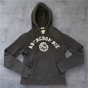 Abercrombie & Fitch hoodie - Mörkgrå hoodie från Abercrombie & Fitch med vit broderad logga och emblem på bröstet. Tröjan har huva med snörning och en stor magficka. Mjuk insida och klassisk collegekänsla, perfekt för chill dagar.