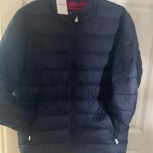 Mörkblå pufferjacka Jack & Jones XL - Snygg mörkblå pufferjacka från Jack & Jones med röd insida. Jackan har diskret logotyp på ärmen, dragkedja framtill och två fickor med dragkedja. Perfekt för kyliga dagar och har en clean, sportig look. Prislappen sitter fortfarande på.