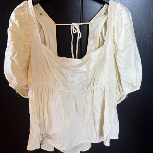 Gul blus med öppen rygg  - Söt gul blus med puffiga korta ärmar och fyrkantig ringning. Toppen har knytband i ryggen och en luftig, flowy passform. Tillverkad i ett lätt linnematerial som ger en avslappnad och trendig vibe. Perfekt för dig som gillar romantiska detaljer och en feminin look. Ryggen är öppen 