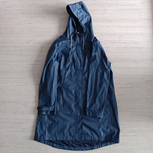 Blå regnjacka från Peter Patter - Snygg marinblå regnjacka från Peter Patter med huva och dragsko. Jackan har knappar framtill, justerbara ärmslut och diskret logga på bröstet och ärmen. Tillverkad i polyester och perfekt för regniga dagar.