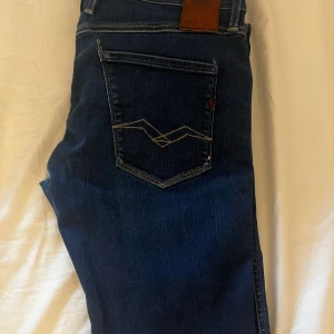 Mörkblå Replay jeans, modell Waitom - Säljer ett par mörkblå jeans från Replay, modell Waitom. Klassisk femficksdesign med snygga kontrastsömmar på bakfickan och läderpatch i midjan. Jeansen är tillverkade i bomull och har en rak passform som funkar till det mesta.