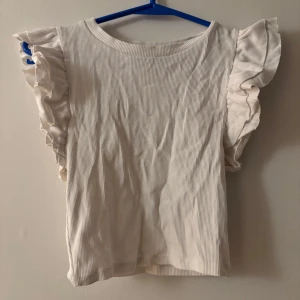 Vit ribbad topp med volangärm från Zara - Supersöt vit ribbad topp från Zara med volangdetaljer på axlarna. Toppen har rund halsringning och är ärmlös med breda volangärmar som ger en extra touch. Perfekt för dig som gillar en feminin och trendig stil.