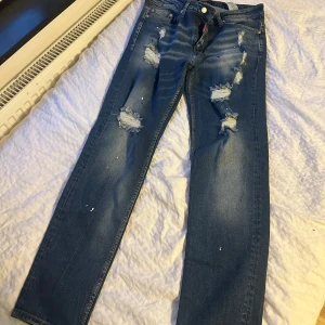 Dsquared2 blå slitna jeans herr - Säljer ett par blå jeans från Dsquared2 med coola slitningar och färgstänk. Jeansen har klassisk femficksdesign, rak passform och tydliga distressed detaljer framtill och baktill. Materialet är jeans i bomull och de har en snygg, avslappnad vibe.