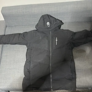 Svart pufferjacka  - Svart pufferjacka från sail racing med huva och hel dragkedja. Jackan har två sidofickor och en bröstficka med dragkedja. Fodrad insida för extra värme och stilren look. Perfekt för kalla dagar och enkel att matcha med allt.