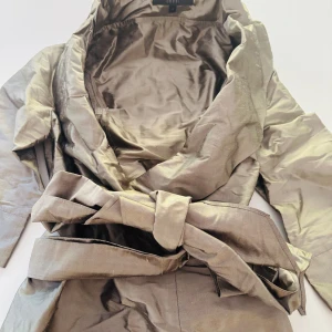 Beige omlottjacka från Coast - Snygg beige omlottjacka från Coast med bred krage och knytskärp i midjan. Jackan har trekvartsärmar och är tillverkad i ett glansigt material som ger en lyxig känsla. Perfekt för dig som vill ha en stilren och trendig look.