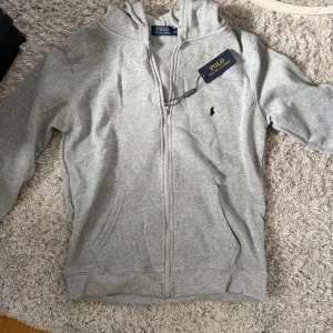 Grå hoodie från Polo Ralph Lauren - Snygg ljusgrå hoodie från Polo Ralph Lauren med dragkedja framtill, huva och klassisk broderad logga på bröstet. Tillverkad i mjuk bomull och har praktiska fickor på magen. Perfekt för en chill och stilren look. Pris kan diskuterats