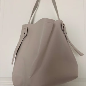 Beige axelväska från H&M i skinnimitation - Stilren beige axelväska från H&M i skinnimitation med bred axelrem och snygga spänndetaljer på sidorna. Väskan har magnetlås i guld och minimalistisk design som passar till många outfits.