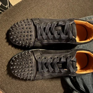Marinblåa Christian Louboutin sneakers med nitar - Säljer ett par svarta sneakers från Christian Louboutin med coola svarta nitar på tån och klassisk röd sula. Skorna har snörning och är gjorda i mocka med läderdetaljer på insidan. Perfekta för dig som vill sticka ut med en edgy vibe. 