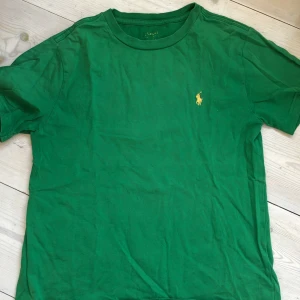 Grön t-shirt från Ralph Lauren, strl 10-12 - Snygg grön t-shirt från Ralph Lauren med klassisk gul broderad logga på bröstet. T-shirten har rund halsringning och korta ärmar. Perfekt basic-plagg med stilren design och skön passform.