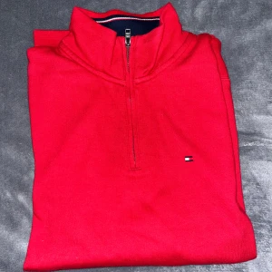 Röd half zip tröja Tommy Hilfiger - Röd långärmad tröja från Tommy Hilfiger med half zip och liten broderad logga på bröstet. Klassisk ståkrage och mjukt bomullsmaterial, perfekt för lager-på-lager. Snygg och enkel att matcha med jeans eller chinos.