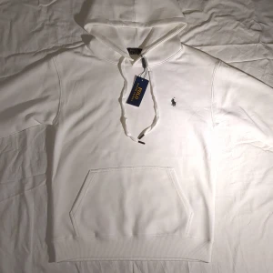 Vit hoodie från Polo Ralph Lauren - Snygg vit hoodie från Polo Ralph Lauren med klassisk broderad logga på bröstet. Tröjan har huva med snörning, känguruficka och ribbade muddar. Perfekt för en clean och stilren look. Tillverkad i mjuk bomull för maximal komfort.