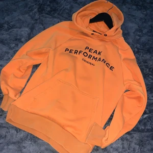 Orange hoodie från Peak Performance - Säljer en snygg orange hoodie från Peak Performance med svart text på bröstet. Tröjan har huva, magficka och ribbade muddar vid ärmslut och nederkant. Perfekt för dig som gillar streetwear och vill sticka ut med färg.