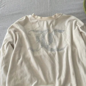 Vit Juicy Couture tröja med strass - Vit långärmad tröja från Juicy Couture med stort glittrigt JC-logo i strass framtill. Mjuk och skön, perfekt för dig som gillar en clean men ändå glam look. Rund halsringning och normal passform.