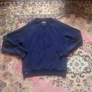 Mörkblå sweatshirt från Morris Gents - Mörkblå sweatshirt från Morris Gents i storlek medium. Tröjan har ett broderat emblem på bröstet med texten 'University of Morris' och klassiska muddar vid ärmslut och nederkant. Skön och stilren modell i bomull, perfekt för lager-på-lager.