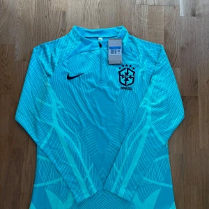 Nike Brasil långärmad fotbollströja M - Snygg långärmad Brasilien fotbollströja från Nike i ljusblått med grafiskt mönster och dragkedja vid halsen. Broderat CBF-märke på bröstet och Nike-logga. Tillverkad i lätt och ventilerande material, perfekt för träning eller match.