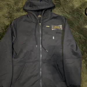 Marinblå hoodie från Polo Ralph Lauren - Snygg marinblå hoodie från Polo Ralph Lauren med dragkedja framtill, huva med snörning och klassisk logga broderad på bröstet. Tröjan har känguruficka och är tillverkad i mjuk bomullsmix, perfekt för chill dagar.