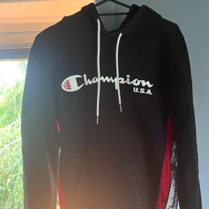 Svart stilren Champion hoodie - Svart hoodie från Champion med stor vit logga och texten U.S.A. på bröstet. Tröjan har vita dragsnören i huvan och röda samt vita paneler på sidorna. Klassisk känguruficka framtill och mjukt material som känns skönt mot huden.