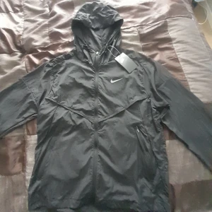 Svart Nike vindjacka med huva M - Svart vindjacka från Nike i storlek M. Jackan har huva, dragkedja framtill och diskret Nike-logga på bröstet. Helt ny från nike jag skulle sälja den för att den vart för liten för mig men den är onvänd
