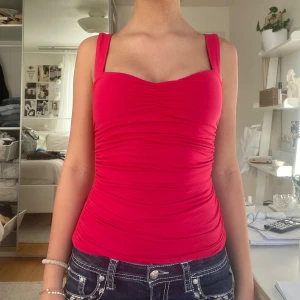 Röd figurnära linnetopp med rynk - Säljer en snygg röd linnetopp med breda axelband och rynkade detaljer framtill. Toppen har en söt sweetheart-urringning och är figurnära i modellen. Perfekt att styla med jeans eller kjol för en trendig look.