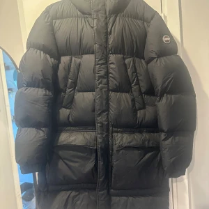 Svart dunjacka från Colmar storlek 54 - Säljer en svart lång dunjacka från Colmar med huva och stora fickor. Jackan har dragkedja och knappar framtill samt en puffig, quiltad design som håller dig varm under kalla dagar. Perfekt för dig som vill ha en stilren och funktionell vinterjacka. Finns ett hål på armen och slitage vid nedsidan av dragkedjan (se sista 2 bilderna) det är inget som syns eller påverkar jackan. Pris kan diskuteras är även öppen för byten mot j.lindeberg parka som tex snowmass betalar mellanskillnad då.