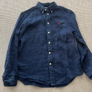 Mörkblå linneskjorta från Ralph lauren - Klassisk mörkblå skjorta från Polo Ralph Lauren med vit knappstängning och röd broderad logga på bröstet. Skjortan har lång ärm, button down-krage och är tillverkad i linnetyg. Perfekt för en clean och stilren look men också för fästligare tillfällen.