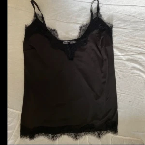 Svart linnetopp med spets från Coster - Svart linnetopp från Coster Copenhagen med tunna axelband och v-ringning. Toppen har snygga spetsdetaljer längs halsringning och nederkant som ger en lyxig känsla. Perfekt att styla med jeans eller kjol för en trendig look.