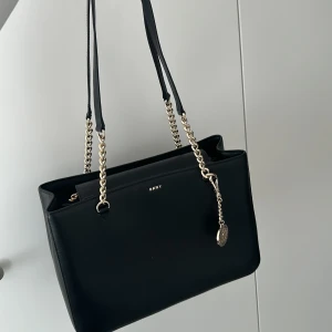 Svart axelväska från DKNY i skinn - Stilren svart axelväska från DKNY i slätt skinn med guldfärgade kedjedetaljer och logotyp framtill. Väskan har rektangulär form, dubbla axelremmar i kedja och läder samt en dekorativ tag med DKNY-logga. Perfekt för dig som gillar klassisk och lyxig look.