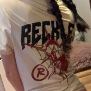 Vit RECKLESS t-shirt med tryck - Vit t-shirt från RECKLESS med stort grafiskt tryck i svart och rött framtill. Klassisk passform med korta ärmar och rund halsringning. Perfekt för dig som gillar streetwear vibes och coola prints.