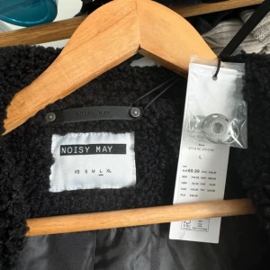 Svart teddyjacka från Noisy May - Mysig svart teddyjacka från Noisy May i storlek L. Jackan har en fluffig och grov yta, klassisk krage och sidofickor. Insidan är fodrad med slätt tyg och jackan stängs med knappar. Perfekt för dig som gillar en chill och trendig look.