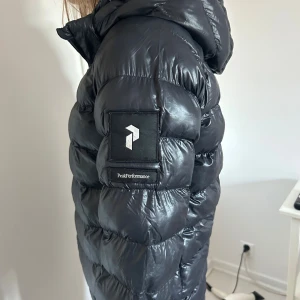 Svart pufferjacka från Peak Performance - Säljer den populära svarta pufferjackan från Peak Performance. Jackan har en dragkedja framtill och en praktisk huva. Perfekt för kyliga dagar med sin vadderade design. Är som en XXS.