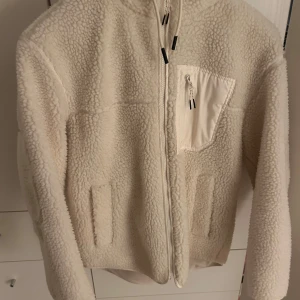 Beige teddyjacka med huva - Mysig beige teddyjacka med huva och hel dragkedja framtill. Jackan har en bröstficka med dragkedja och två sidofickor. Perfekt för kalla dagar och har en riktigt chill vibe. Passar på xs-m
