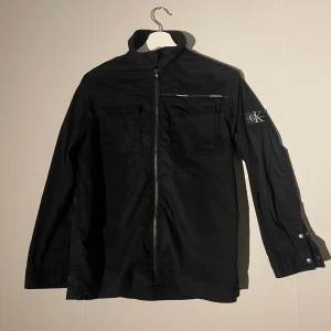 Svart overshirt från Calvin Klein Jeans - Svart overshirt från Calvin Klein Jeans med dragkedja framtill, två bröstfickor och logotyp-patch på armen. Perfekt för stilren look.