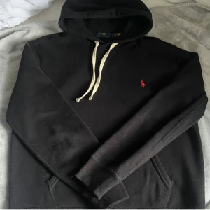 Svart hoodie från Polo Ralph Lauren - Svart hoodie från Polo Ralph Lauren med vit snörning och klassisk röd logga broderad på bröstet. Tröjan har huva, känguruficka och långa ärmar. Perfekt för en chill och stilren look.