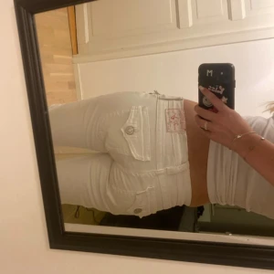 Vita True Religion jeans, bootcut, stl 27 - Säljer ett par vita jeans från True Religion i klassisk bootcut-modell. Byxorna har snygga fickdetaljer med lock och knappar bak, samt ikonisk True Religion-lapp. Tillverkade i bomull med en skön passform och låg midja. Perfekta för dig som vill sticka ut.