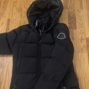 Svart Moncler dunjacka med huva - Säljer en svart dunjacka från Moncler med huva och dragkedja. Jackan har Moncler-logga på ärmen och tryckt Moncler-text på insidan av huvan. Puffig modell med två sidofickor med dragkedja. Perfekt för kalla dagar och riktigt snygg streetstil.