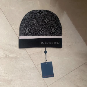 Svart och grå Louis Vuitton mössa - Exklusiv svart och grå mössa från Louis Vuitton med klassiskt monogrammönster och broderad logga framtill. Mössan har en bred, ljusgrå kant och är tillverkad i ett mjukt, stickat material som känns lyxigt och bekvämt. Perfekt för dig som vill ha en stilren accessoar med premiumkänsla.