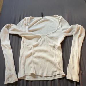 Vit ribbad långärmad topp Divided - Säljer en vit ribbad långärmad topp från Divided. Toppen har en snygg sömdetalj framtill som ger en modern look och en bred, fyrkantig urringning. Materialet är stretchigt och sitter tajt mot kroppen. Perfekt att styla med jeans.